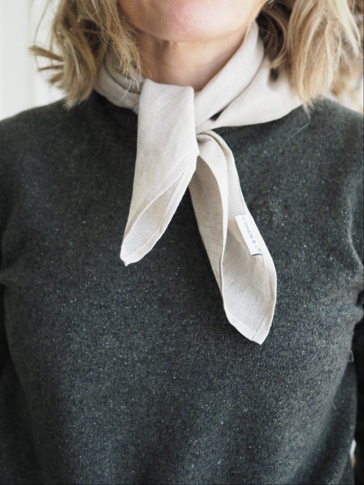 Woman wearing a dark gray sweater with a beige scarf around her neck against a neutral background